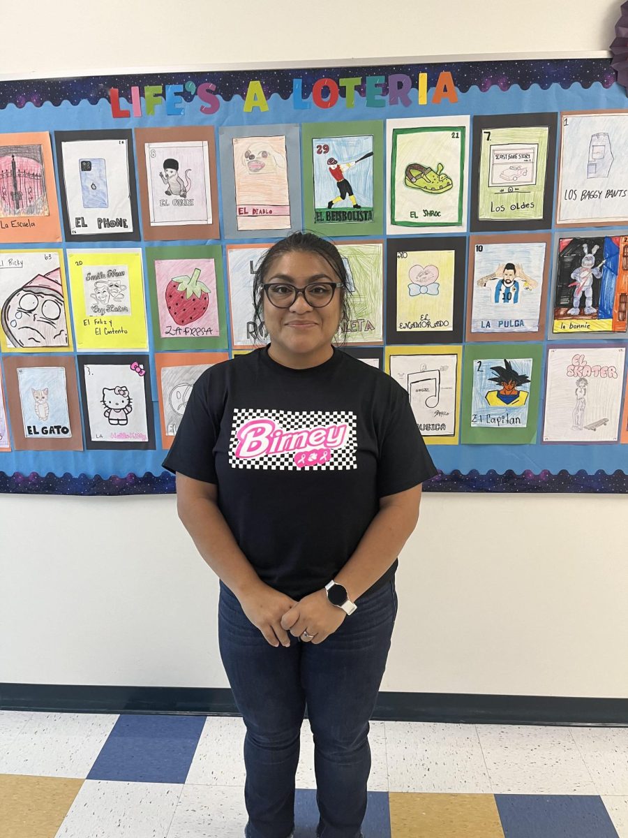 This is Ms. Rodriguez outside of her classroom next to her bulletin board which has pictures of Loteria cards that her students drew. 